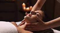 A close-up shot of a woman with her eyes closed receiving a relaxing facial massage from a spa therapist in a dimly lit room with glowing candles in the background.